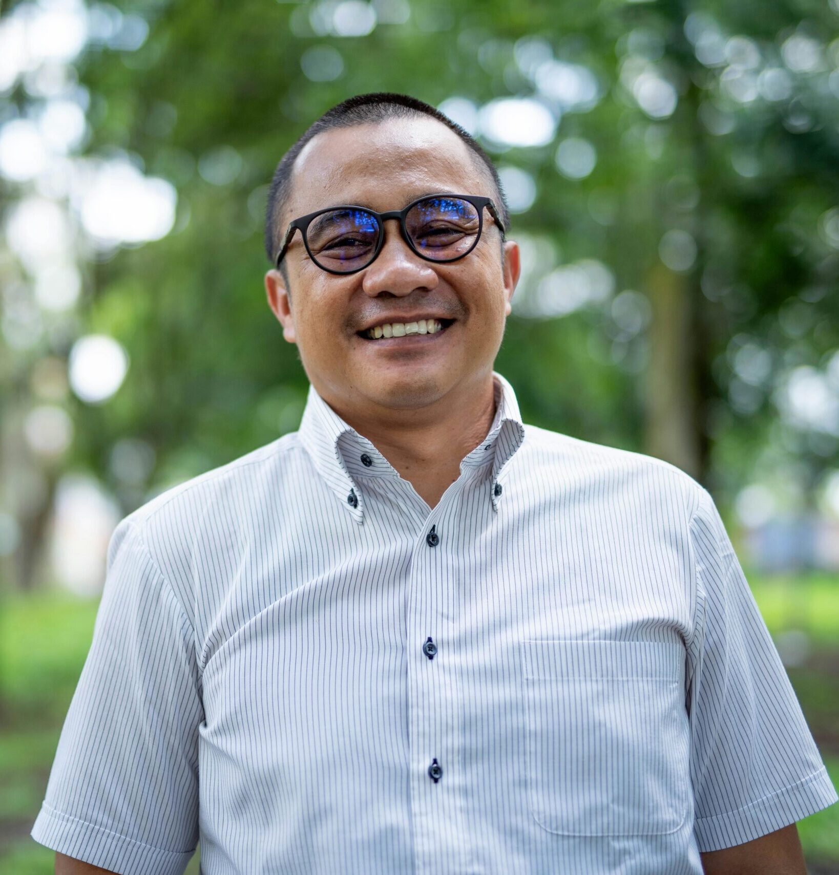 Portrait of a smiling Asian man with eyeglasses standing in a green park setting.